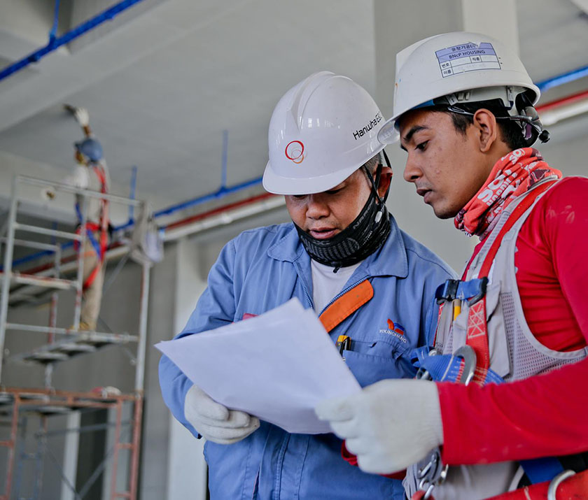 Two men in hard hats and safety vests reviewing paperwork at a construction site.