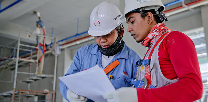 Two men in hard hats and safety vests reviewing paperwork at a construction site.